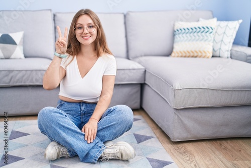 Young caucasian woman sitting on the floor at the living room showing and pointing up with fingers number two while smiling confident and happy.