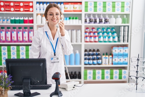 Young blonde woman pharmacist talking on telephone using computer at pharmacy