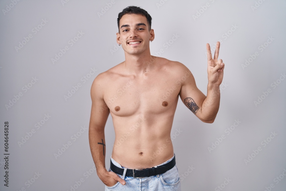 Handsome hispanic man standing shirtless smiling looking to the camera showing fingers doing victory sign. number two.