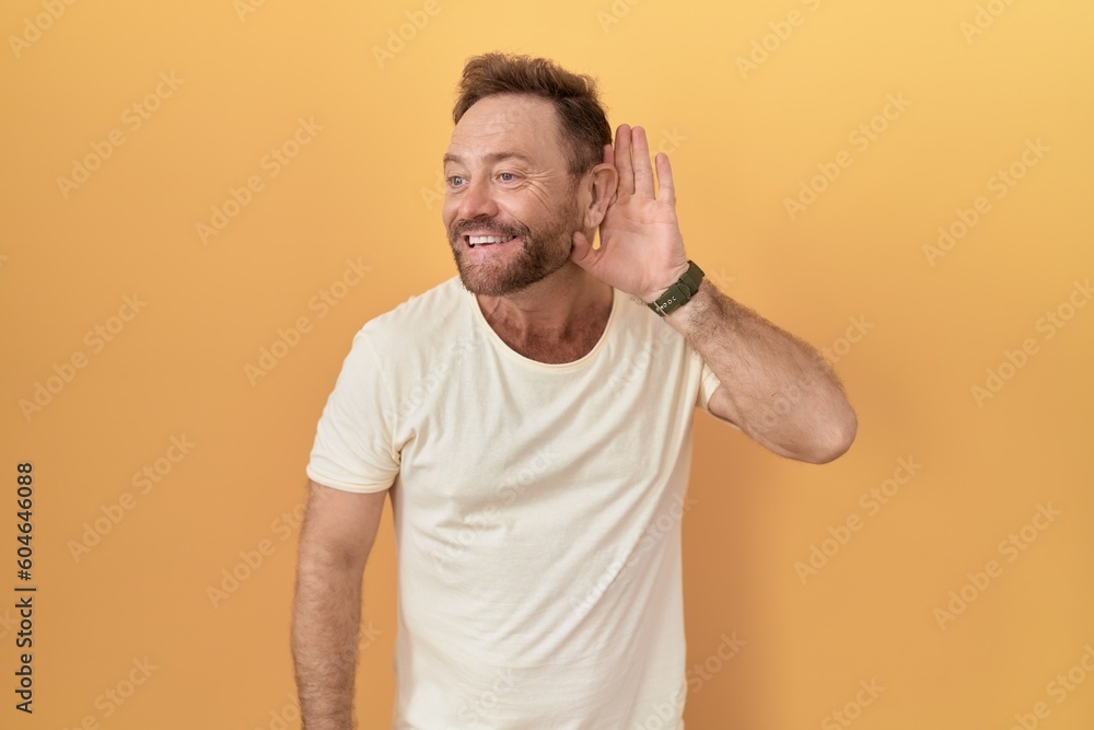 Middle age man with beard standing over yellow background smiling with hand over ear listening an hearing to rumor or gossip. deafness concept. Middle age man with beard standing over yellow background smiling with hand over ear listening an hearing to rumor or gossip. deafness concept.