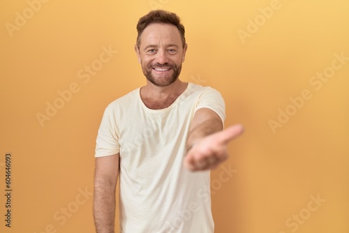 Middle age man with beard standing over yellow background smiling cheerful offering palm hand giving assistance and acceptance. © Krakenimages.com