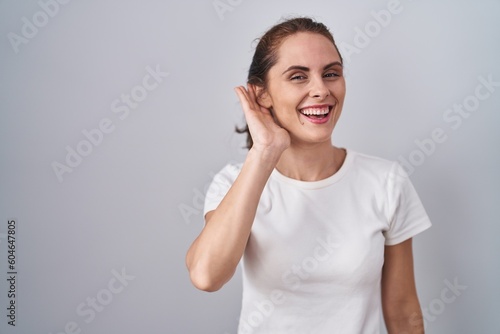 Beautiful brunette woman standing over isolated background smiling with hand over ear listening an hearing to rumor or gossip. deafness concept. © Krakenimages.com