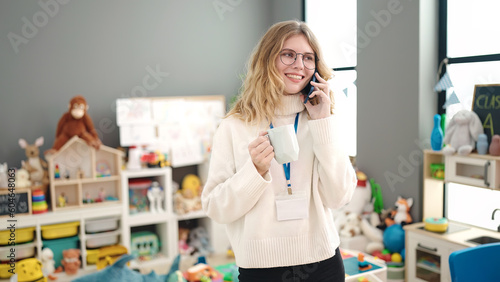 Young blonde woman preschool teacher talking on smartphone drinking coffee at kindergarten