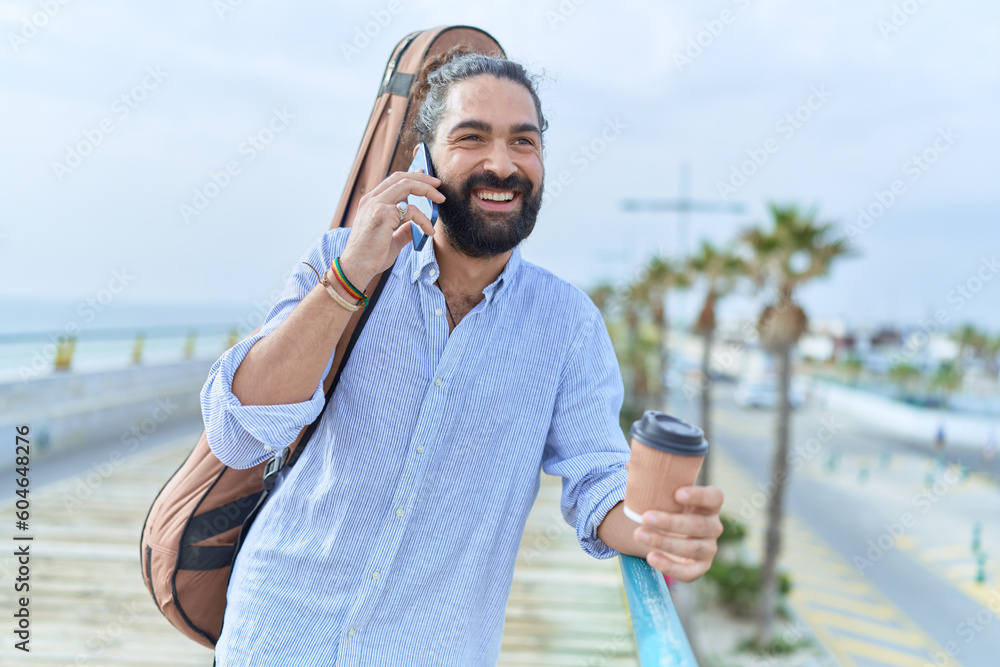 Young hispanic man musician talking on smartphone drinking coffee at seaside