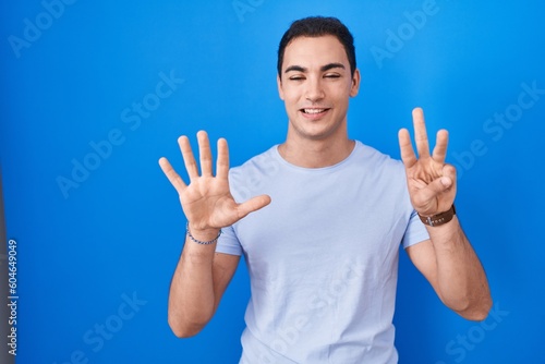 Young hispanic man standing over blue background showing and pointing up with fingers number eight while smiling confident and happy.
