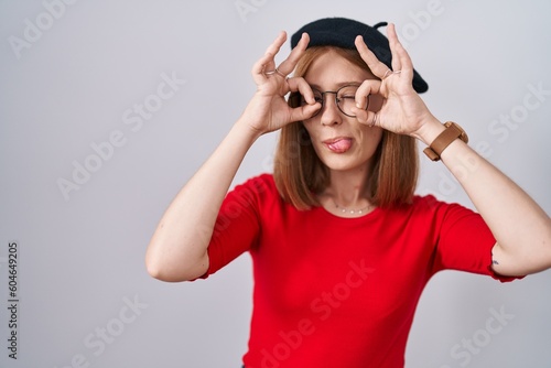 Young redhead woman standing wearing glasses and beret doing ok gesture like binoculars sticking tongue out, eyes looking through fingers. crazy expression.