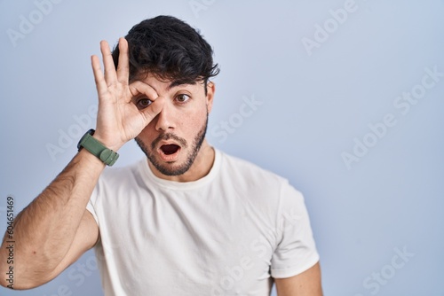 Hispanic man with beard standing over white background doing ok gesture shocked with surprised face, eye looking through fingers. unbelieving expression.