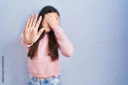 Young brunette woman standing over blue background covering eyes with hands and doing stop gesture with sad and fear expression. embarrassed and negative concept.