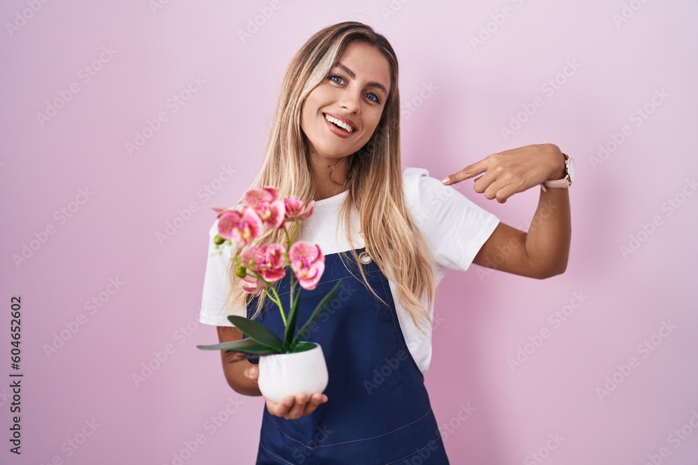 Young blonde woman wearing gardener apron holding plant looking confident with smile on face, pointing oneself with fingers proud and happy. Young blonde woman wearing gardener apron holding plant looking confident with smile on face, pointing oneself with fingers proud and happy.