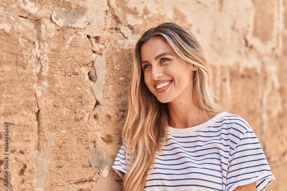 Young blonde woman smiling confident looking to the side over isolated stone background