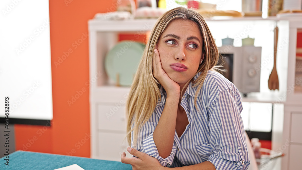 Young blonde woman sitting on table with sad expression at dinning room