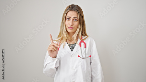 Young blonde woman doctor standing with serious expression saying no with finger over isolated white background