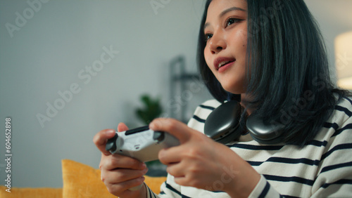 Excited beautiful Asian woman using joystick wireless controller enjoy playing fighting console video game to win highest victory. Girl Gamer sit on sofa in modern living room at home