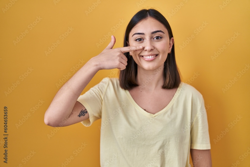 Hispanic girl wearing casual t shirt over yellow background pointing with hand finger to face and nose, smiling cheerful. beauty concept Hispanic girl wearing casual t shirt over yellow background pointing with hand finger to face and nose, smiling cheerful. beauty concept