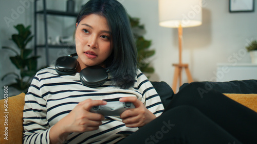 Excited beautiful Asian woman using joystick wireless controller enjoy playing fighting console video game to win highest victory. Girl Gamer sit on sofa in modern living room at home