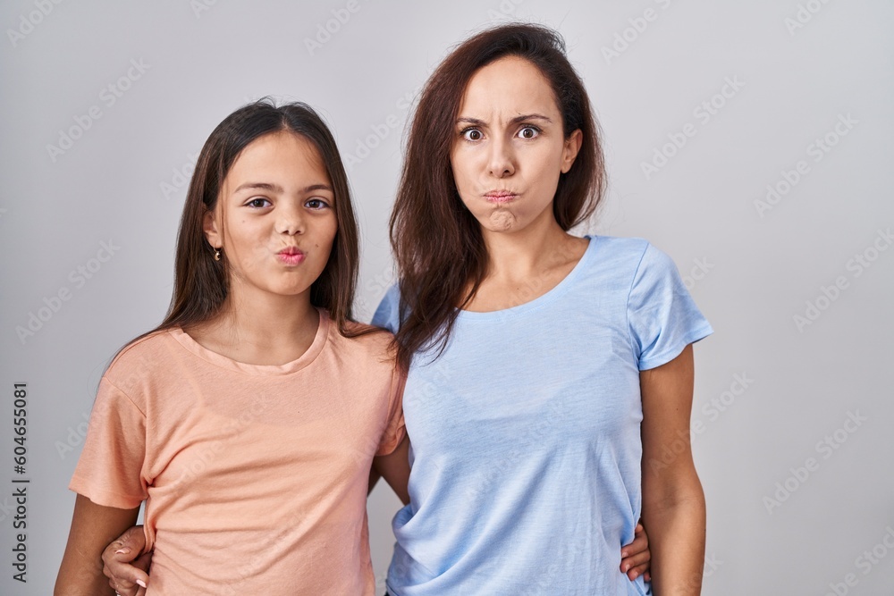 Young mother and daughter standing over white background puffing cheeks with funny face. mouth inflated with air, crazy expression. Young mother and daughter standing over white background puffing cheeks with funny face. mouth inflated with air, crazy expression.