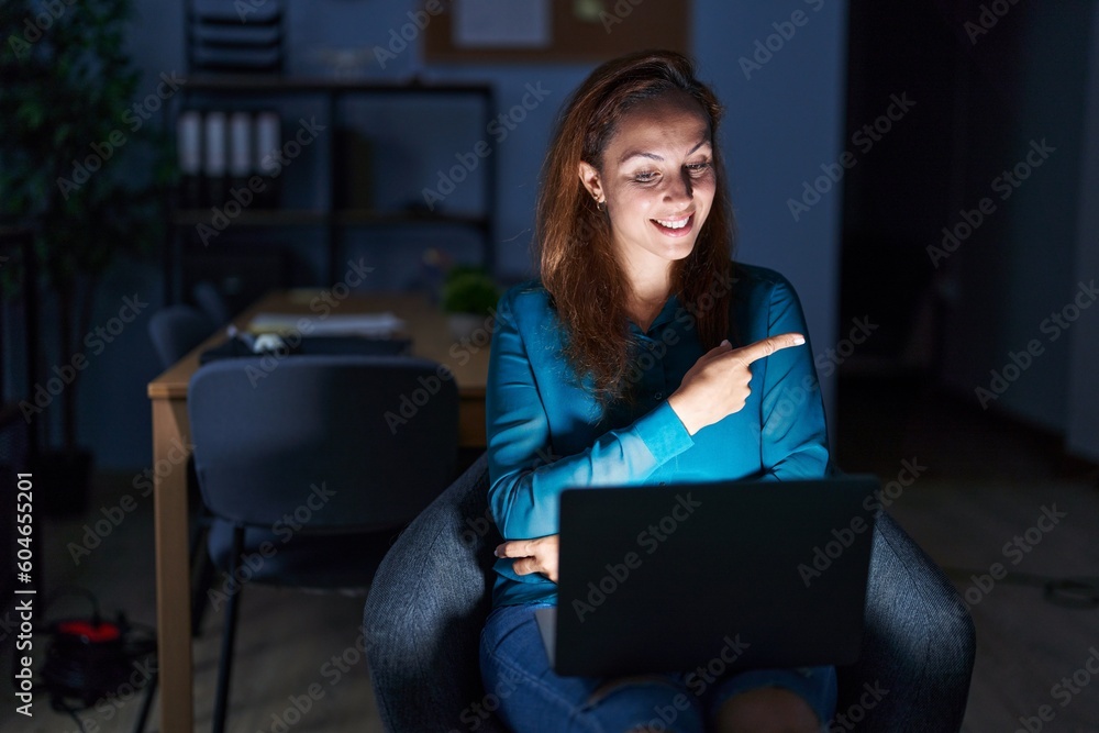 Brunette woman working at the office at night with a big smile on face, pointing with hand and finger to the side looking at the camera. Brunette woman working at the office at night with a big smile on face, pointing with hand and finger to the side looking at the camera.