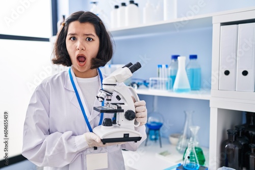 Young hispanic woman working at scientist laboratory holding microscope in shock face  looking skeptical and sarcastic  surprised with open mouth