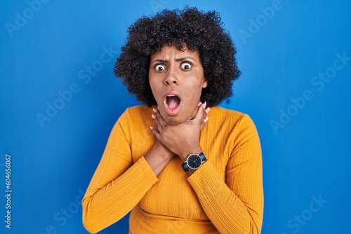 Black woman with curly hair standing over blue background shouting suffocate because painful strangle. health problem. asphyxiate and suicide concept.