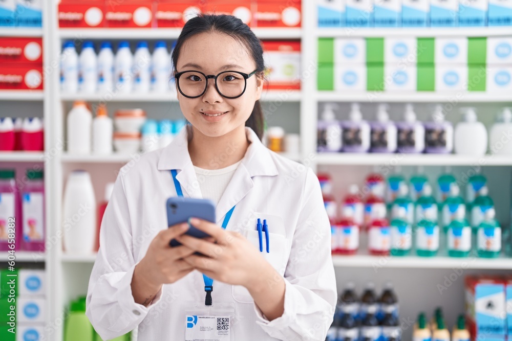 Young chinese woman pharmacist using smartphone working at pharmacy