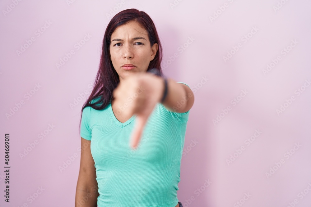Young hispanic woman standing over pink background looking unhappy and angry showing rejection and negative with thumbs down gesture. bad expression. Young hispanic woman standing over pink background looking unhappy and angry showing rejection and negative with thumbs down gesture. bad expression.