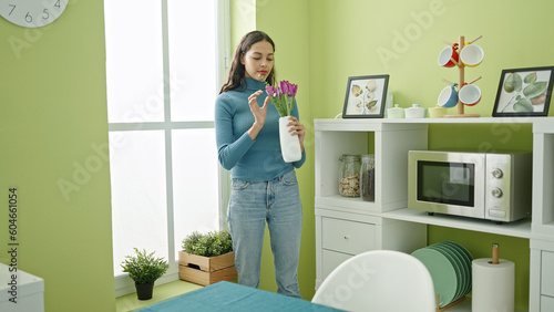 Young beautiful hispanic woman holding flowers in a vase at home
