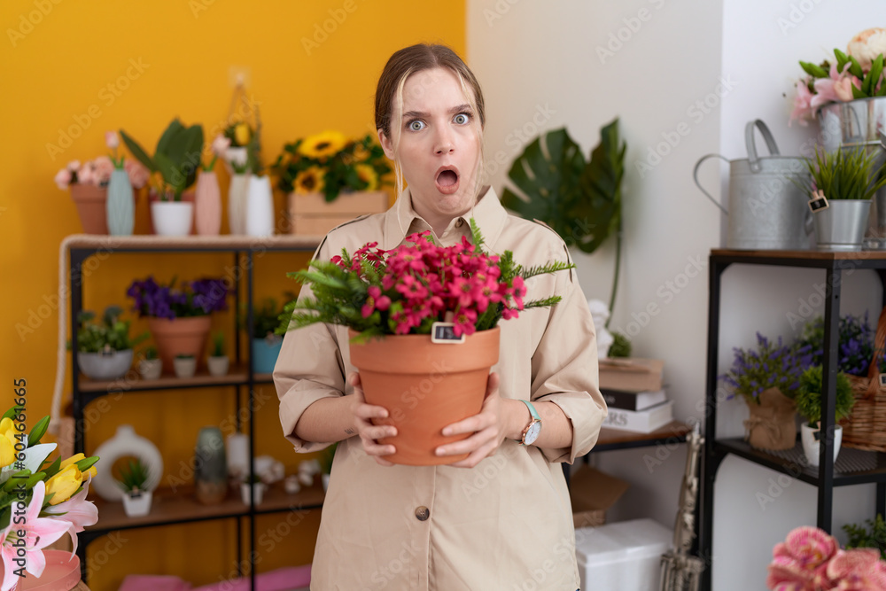 Young caucasian woman working at florist shop holding plant pot in shock face, looking skeptical and sarcastic, surprised with open mouth