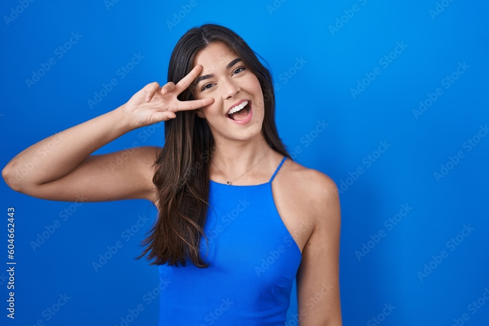 Hispanic woman standing over blue background doing peace symbol with fingers over face, smiling cheerful showing victory Hispanic woman standing over blue background doing peace symbol with fingers over face, smiling cheerful showing victory