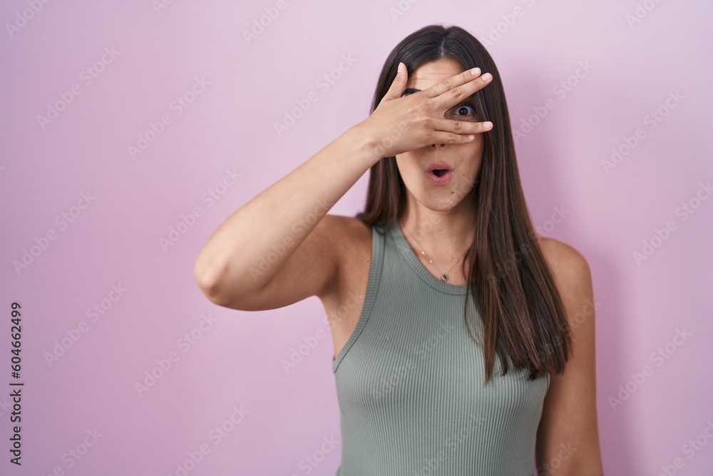 Hispanic woman standing over pink background peeking in shock covering face and eyes with hand, looking through fingers with embarrassed expression. Hispanic woman standing over pink background peeking in shock covering face and eyes with hand, looking through fingers with embarrassed expression.