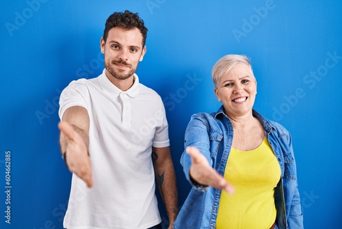 Young brazilian mother and son standing over blue background smiling friendly offering handshake as greeting and welcoming. successful business.