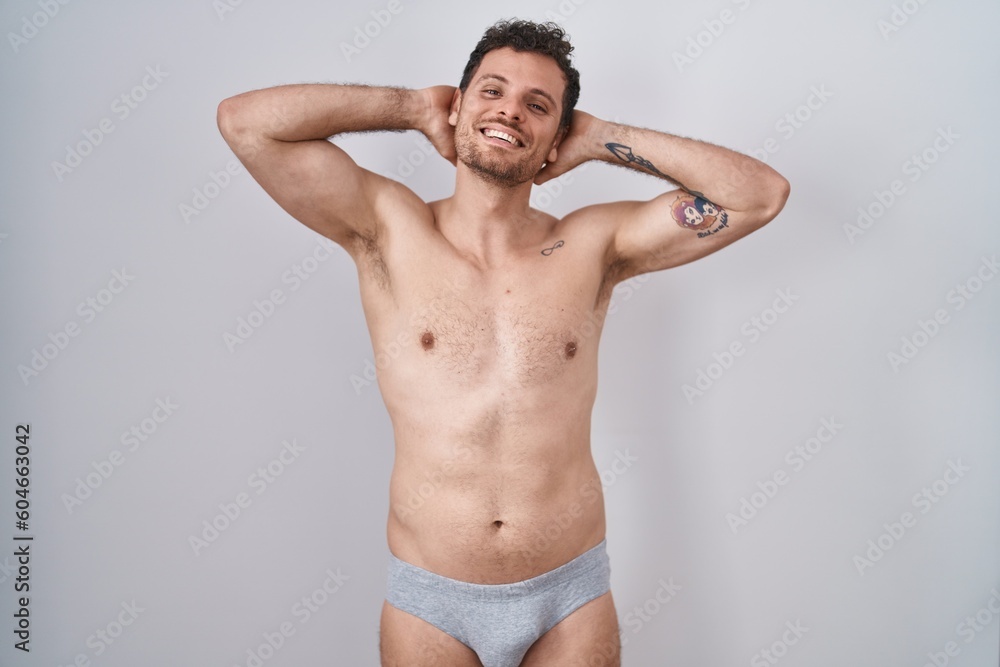 Young hispanic man standing shirtless wearing underware relaxing and stretching, arms and hands behind head and neck smiling happy Young hispanic man standing shirtless wearing underware relaxing and stretching, arms and hands behind head and neck smiling happy
