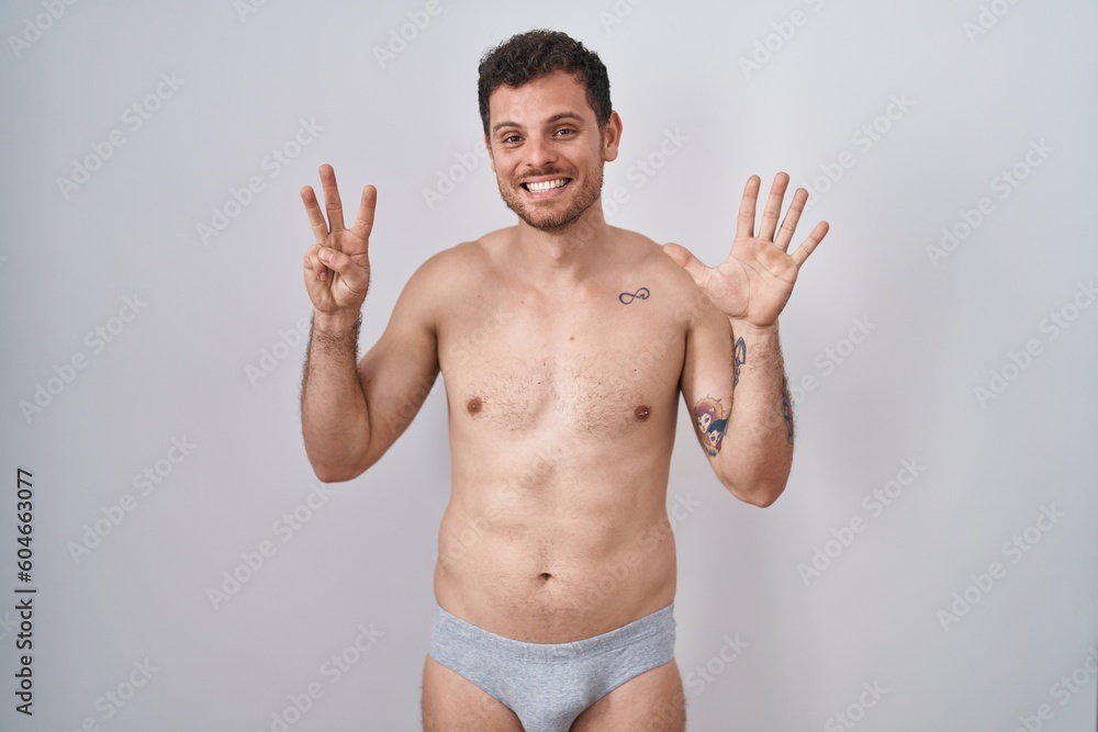 Young hispanic man standing shirtless wearing underware showing and pointing up with fingers number eight while smiling confident and happy. Young hispanic man standing shirtless wearing underware showing and pointing up with fingers number eight while smiling confident and happy.