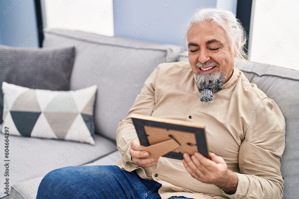 Middle age grey-haired man looking picture sitting on sofa at home Middle age grey-haired man looking picture sitting on sofa at home
