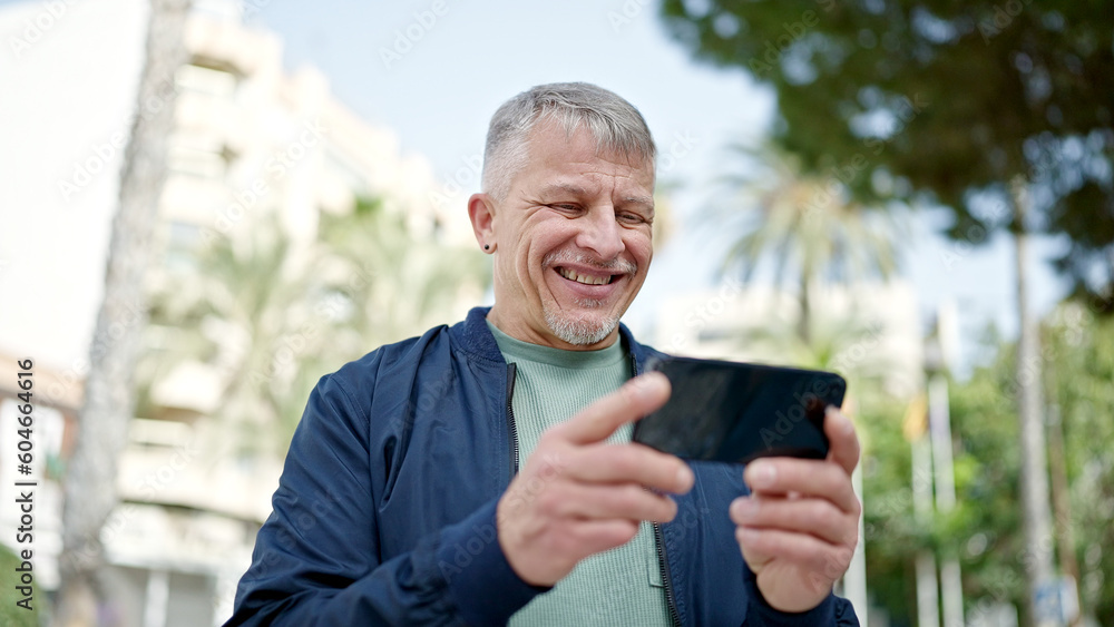 Middle age grey-haired man smiling confident watching video on smartphone at park Middle age grey-haired man smiling confident watching video on smartphone at park