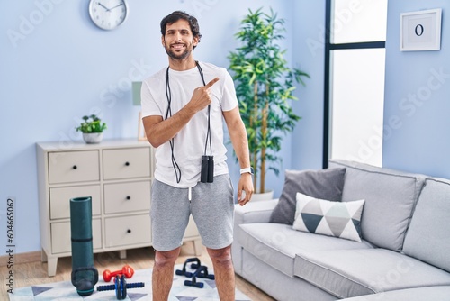 Handsome latin man wearing sportswear at home cheerful with a smile of face pointing with hand and finger up to the side with happy and natural expression on face