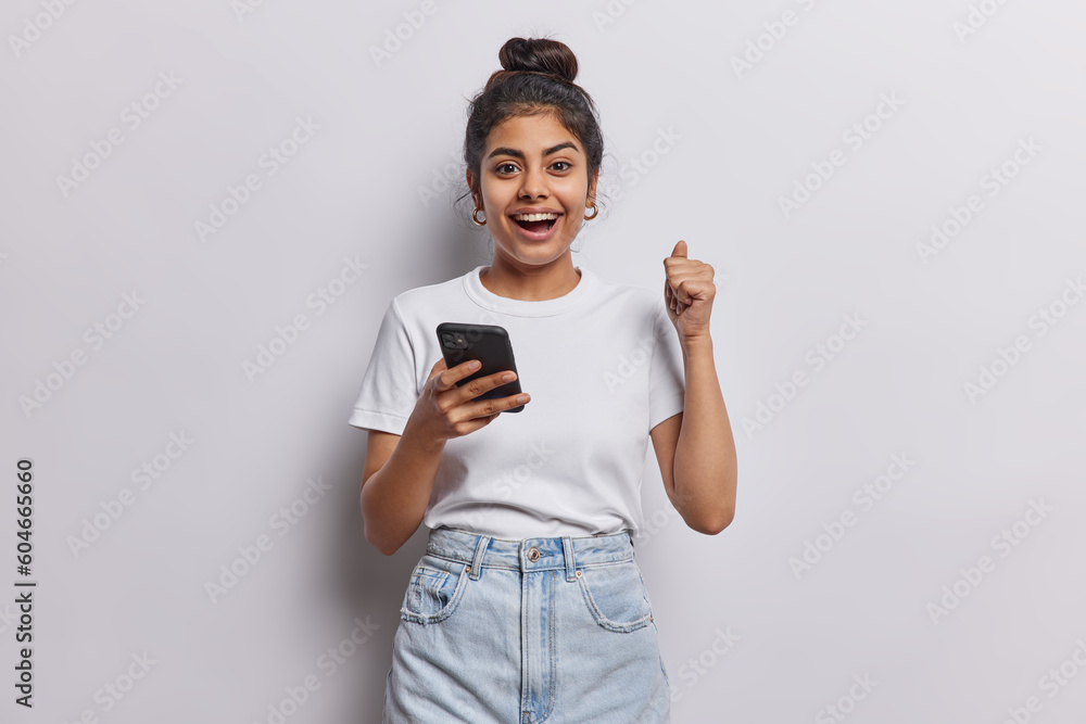 Happy cheerful Indian girl with dark hair combed in bun clenches fist celebrates success holds smartphone dressed in casual basic t shirt jeans isolated over white wall receives message about reward Happy cheerful Indian girl with dark hair combed in bun clenches fist celebrates success holds smartphone dressed in casual basic t shirt jeans isolated over white wall receives message about reward
