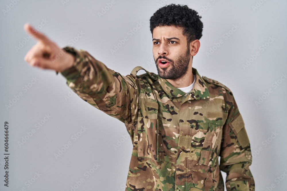 Arab man wearing camouflage army uniform pointing with finger surprised ahead, open mouth amazed expression, something on the front