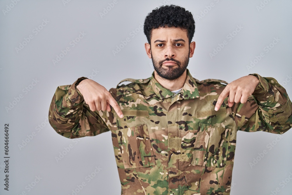 Arab man wearing camouflage army uniform pointing down looking sad and upset, indicating direction with fingers, unhappy and depressed.