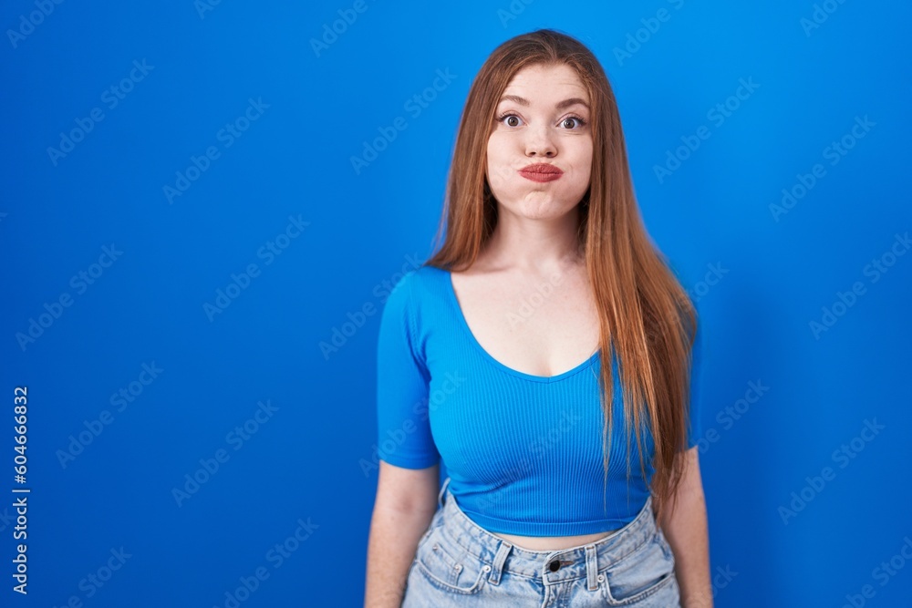 Redhead woman standing over blue background puffing cheeks with funny face. mouth inflated with air, crazy expression. Redhead woman standing over blue background puffing cheeks with funny face. mouth inflated with air, crazy expression.