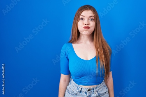 Redhead woman standing over blue background puffing cheeks with funny face. mouth inflated with air, crazy expression.