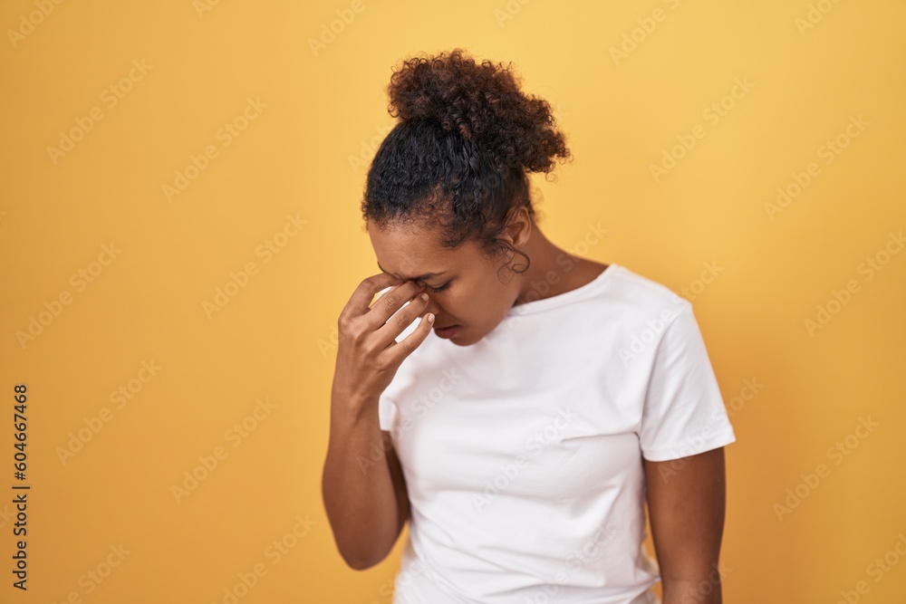 Young hispanic woman with curly hair standing over yellow background tired rubbing nose and eyes feeling fatigue and headache. stress and frustration concept. Young hispanic woman with curly hair standing over yellow background tired rubbing nose and eyes feeling fatigue and headache. stress and frustration concept.