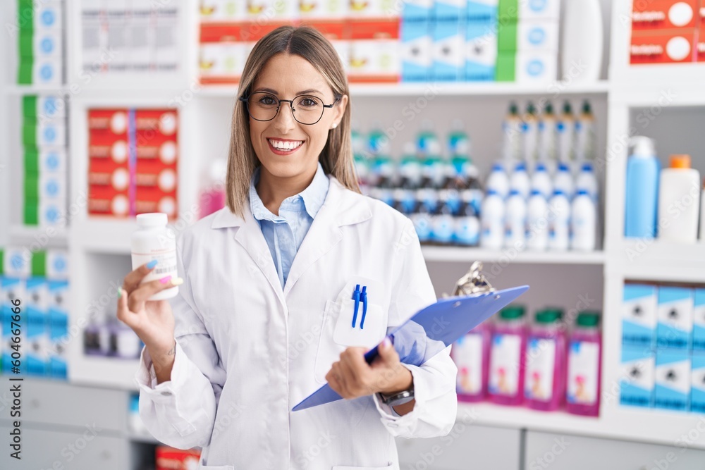 Young beautiful hispanic woman pharmacist holding pills bottle reading document at pharmacy