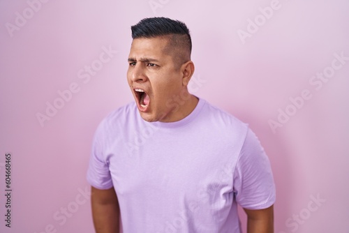 Young hispanic man standing over pink background angry and mad screaming frustrated and furious, shouting with anger. rage and aggressive concept.