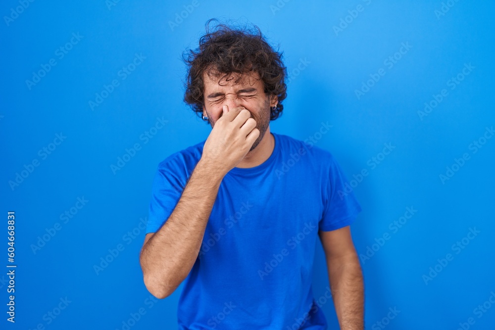 Hispanic young man standing over blue background smelling something stinky and disgusting, intolerable smell, holding breath with fingers on nose. bad smell Hispanic young man standing over blue background smelling something stinky and disgusting, intolerable smell, holding breath with fingers on nose. bad smell