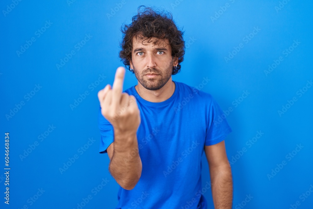 Hispanic young man standing over blue background showing middle finger, impolite and rude fuck off expression Hispanic young man standing over blue background showing middle finger, impolite and rude fuck off expression
