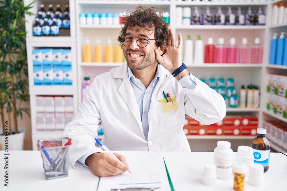 Hispanic young man working at pharmacy drugstore smiling with hand over ear listening an hearing to rumor or gossip. deafness concept.