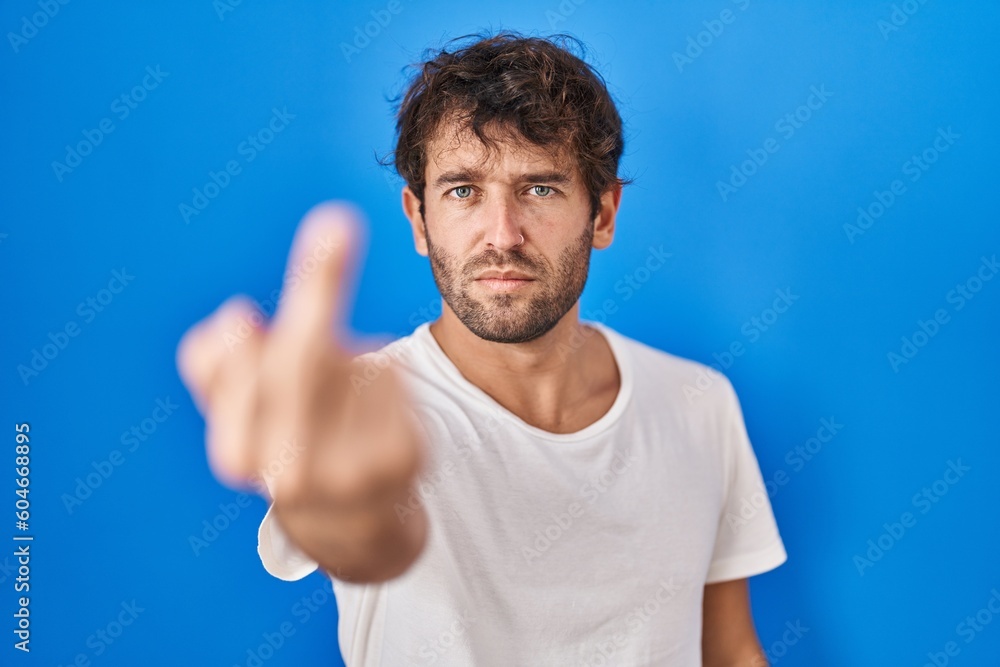 Hispanic young man standing over blue background showing middle finger, impolite and rude fuck off expression Hispanic young man standing over blue background showing middle finger, impolite and rude fuck off expression