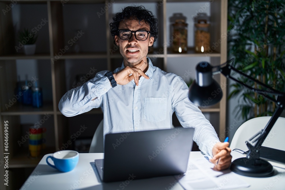 Obraz premium Hispanic man working at the office at night cutting throat with hand as knife, threaten aggression with furious violence