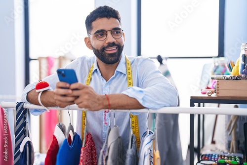 Young arab man tailor using smartphone leaning on clothes rack at tailor shop