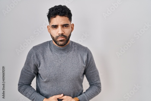 Hispanic man with beard standing over white background with hand on stomach because indigestion, painful illness feeling unwell. ache concept.